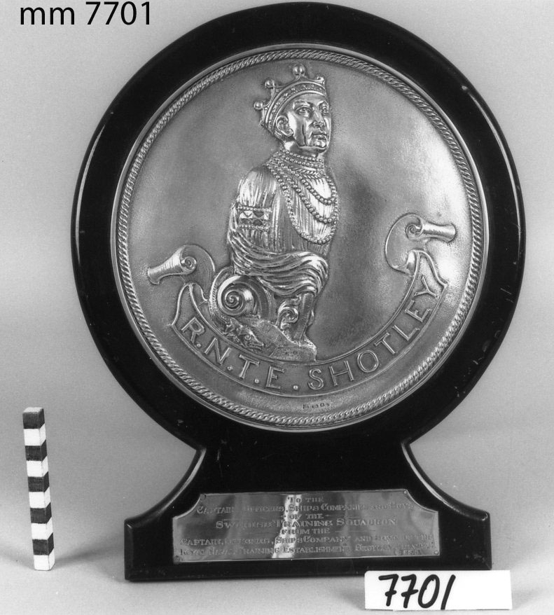 Plakett av silver, fast vid rund träplatta med fot och
stöd av trä, svartlackerat.
Utgör hederspris i idrott, erövrat av en skeppsgosseavdelning vid dess besök i Shotley, England, år 1925.

Medaljongen försedd med kunglig krona och texten:
R. N. T. E.  SHOTLEY.
Inskription på plåt av silver på foten:
To The Captains, Officers, Ships Companies And Boys Of The "Swedish Training Squadron" From The ..... (=upprepning av grader på officerare och manskap från Shotley-laget) July 1925.