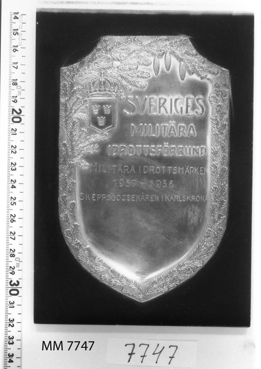 Plakett av silver på platta av trä, svart.
Inskription: Sveriges Militära Idrottsförbund.
Militära Idrottsmärken 1937-1938. Skeppsgossekåren i Karlskrona.