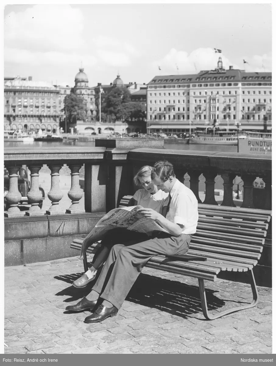 Stockholm. Ett par läser morgontidningen "Stockholm-tidning" på en bänk. I bakgrunden Grand Hotel.