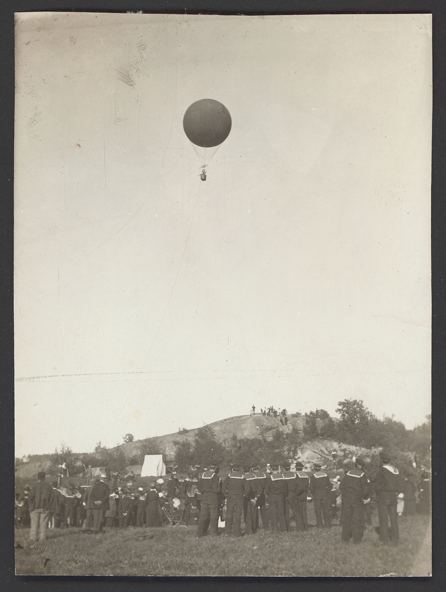 Bilden visar en vätgasballong som stiger upp över en kulle på Vaxö. På marken syns en mängd flottister i sjömanskostymer.