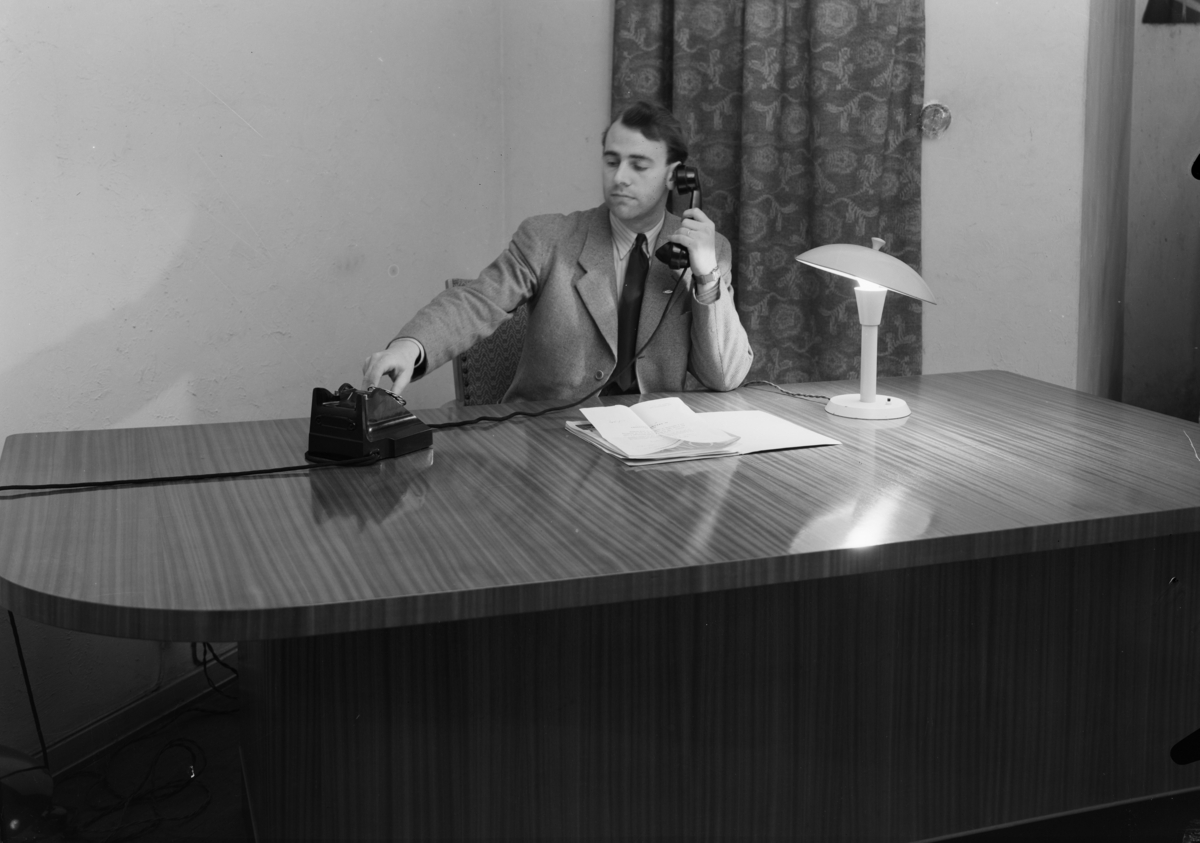 Produktfotografi, P.C.P. bordlampe. En mann sitter ved et stort kontorskrivebord. Han snakker i telefon og har en bunke papirer foran seg. I bakgrunnen henger det et blonstrete draperi. Publisert i Bonytt, mars 1949.