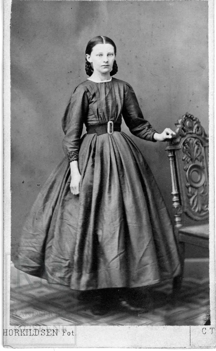 En kvinne med flettet hår iført lang vid kjole med bredt belte. Hun står med hånden hvilende på en utskåret stol.