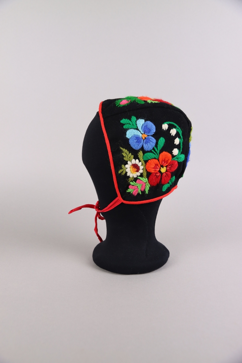 Hätta till flicka av svart kläde med röda band. Hättan är broderad med blomstermotiv av ullgarn i flera färger med påsöm i Dala-Floda-stil.

