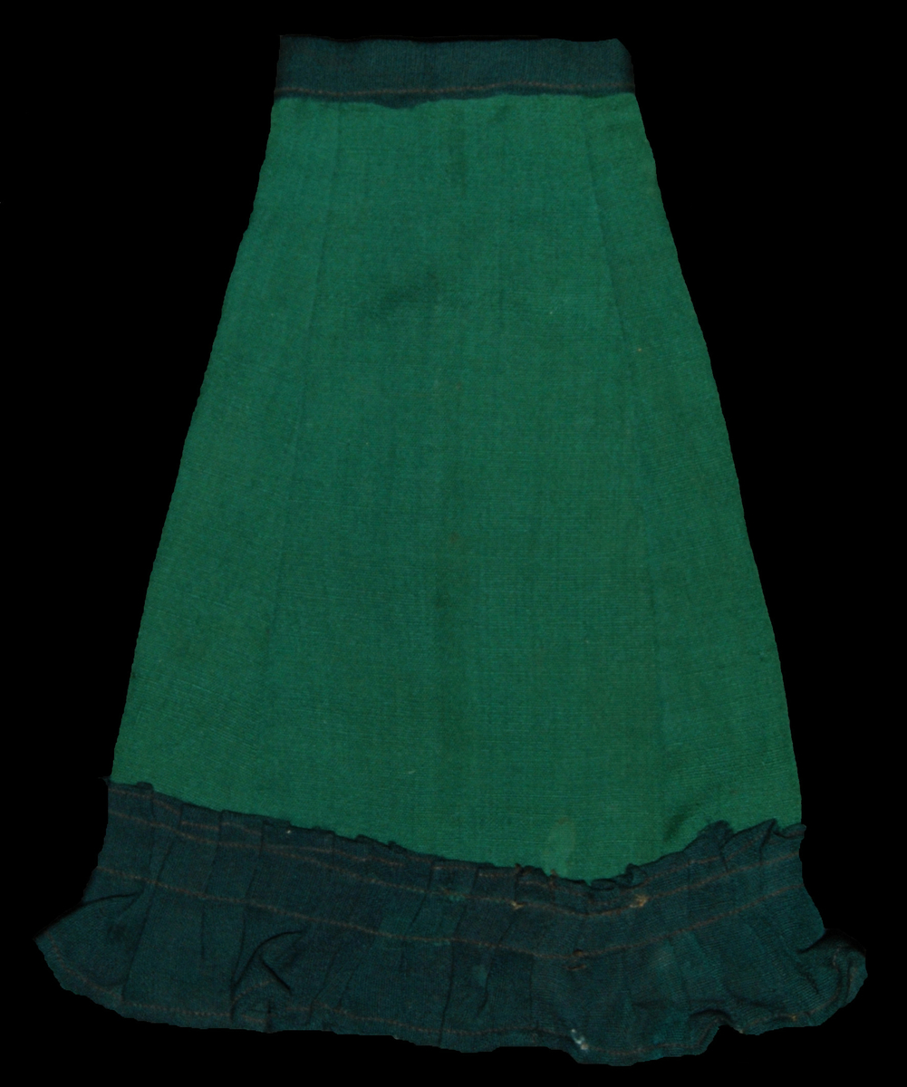 Klänning bestående av grön lång kjol med mörkare grön linning. En bred volangkant av samma mörka tyg som i övre delen av kjolen. Västjacka med spets i halsen. stängs med hyska och hake.
