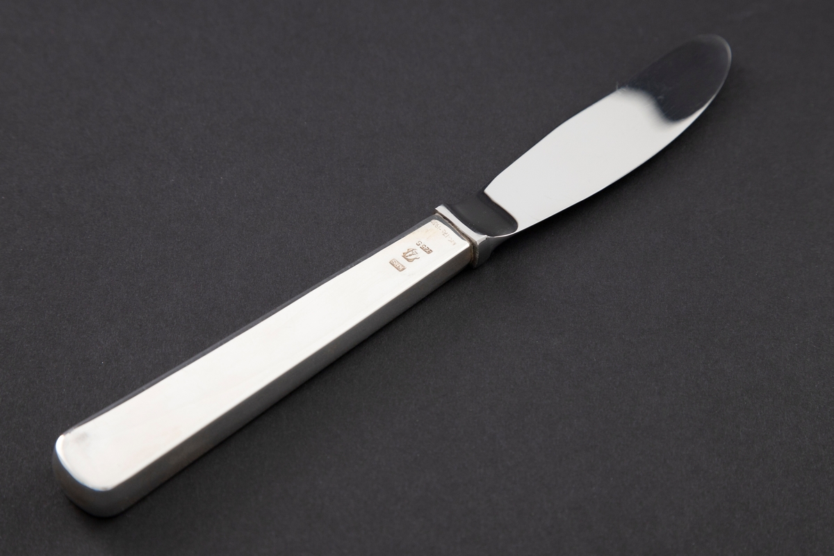 Dessertkniv med skaft i sølv. En rektangulær holk markerer overgangen mellom det lange, tykke skaftet og bladet. Sistnevnte er utført i rustfritt stål. Glatt udekorert overflate.