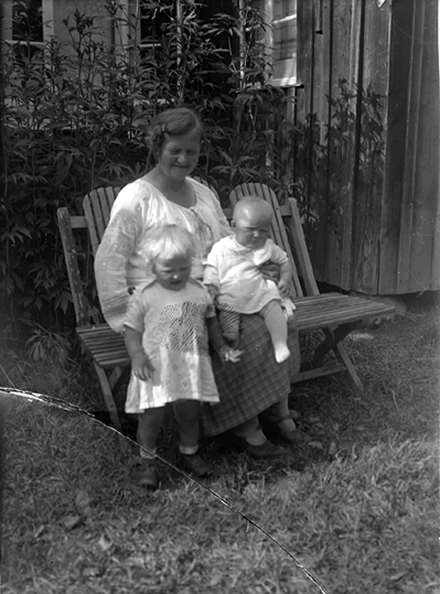 Kvinna med två barn, sitter på trädgårdssoffa framför husvägg. Västerås.