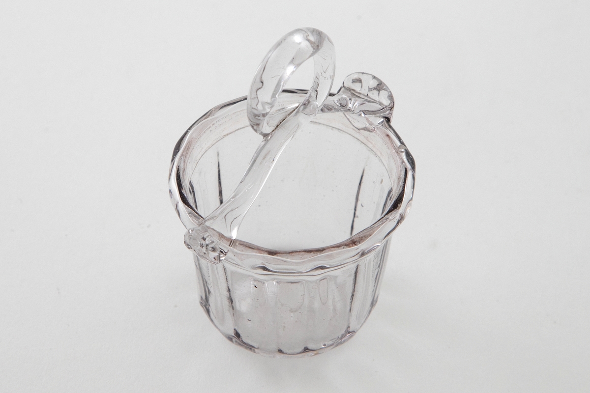 Kurvformet skål i klart glass med en grålig tone. Sirkulært korpus med vertikale riller. En svakt bøyd glasstav med løkkke fungerer som hank. Puntemerke på undersiden av skålen.