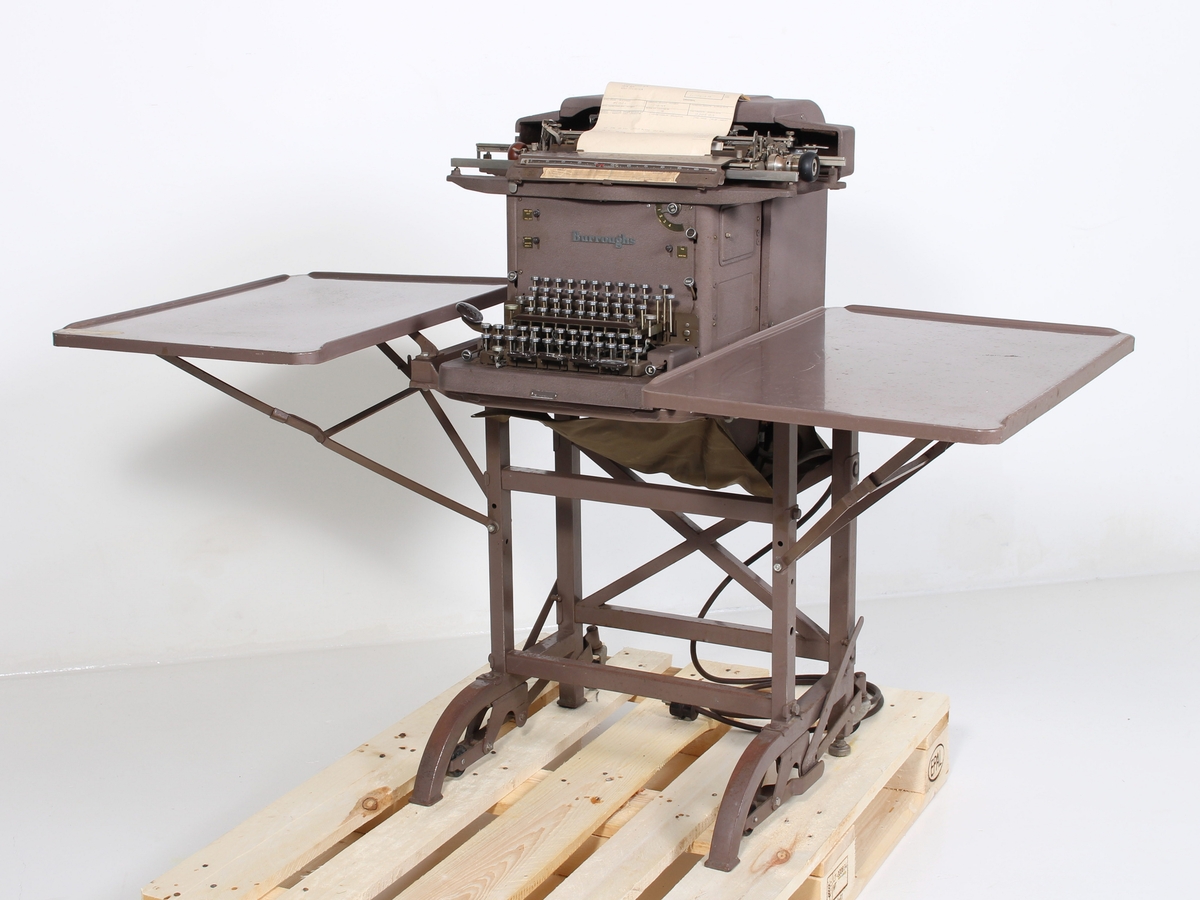 Kombinert regne- og skrivemaskin. Montert på stålrørsstativ med hjul og sammenleggbare bordplater. 
En uferdig faktura sitter igjen i maskinen - denne er datert 12. mars 1985 og avsender er merket Øglænd Metallgruppen.