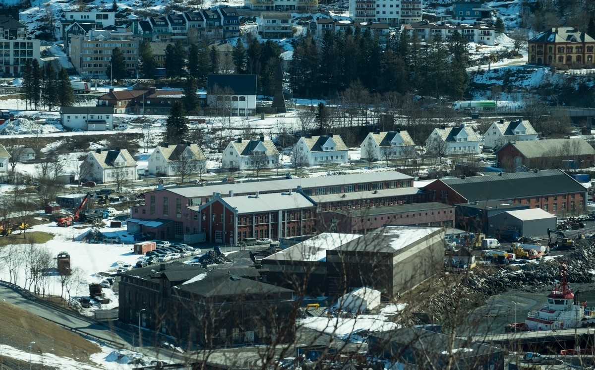 Tidligere LKABs verksteder på havna i Narvik. Hvitmalt husrekke bak er tidligere boliger for adm-ansatte i LKAB, Havnegata 10-22. Sjømannskirka i Narvik, fra baksiden, sees i bakgrunnen . Foto 10. april 2019.