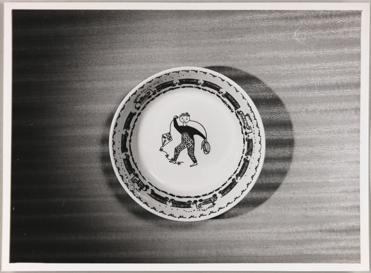 Dyp tallerken tilhørende et barneservise fra Stavangerflint A/S. Motivet i midten er en gutt som flyr drage, og borddekorens motiv er et tog.