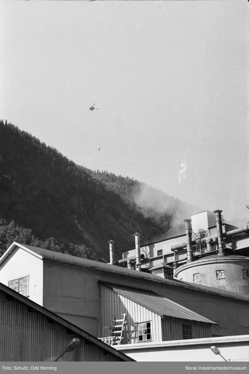 Helikopter med last hengende under i ferdsel over fabrikkområde.