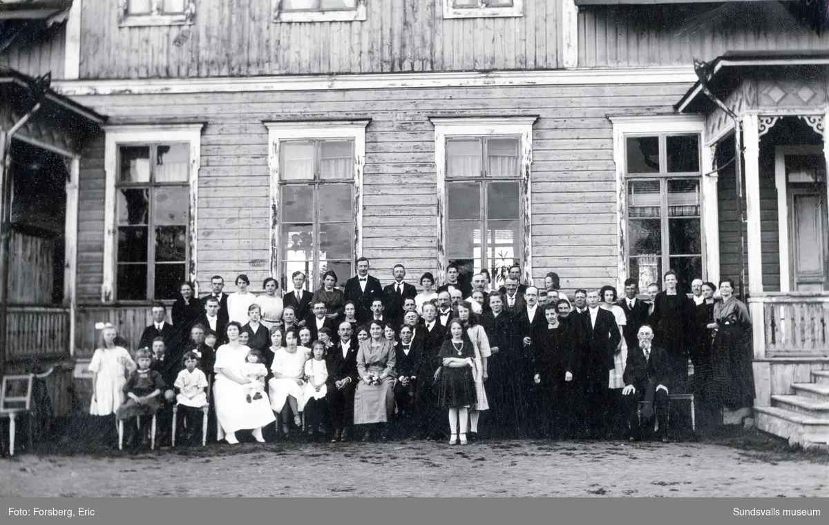 Gruppbild med drygt 60 män, kvinnor och barn framför en äldre träbyggnad. Släktfoto? Ur ett album som tillhört Karin Andersson, Skön.