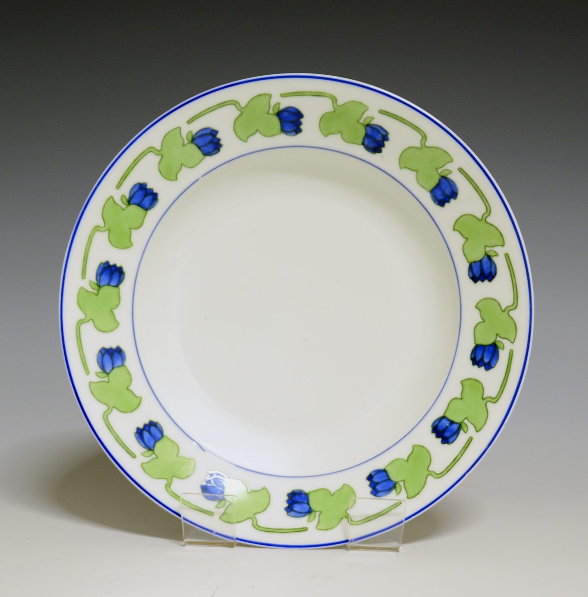 Dyp tallerken i porselen. Hvit glasur. Stilisert bord med blå blomst og grønt blad rundt  fanen.
Modellnr: 15.2
