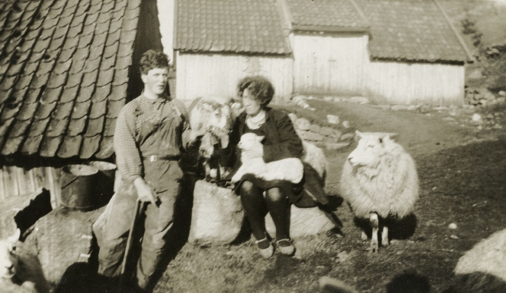Ein ung mann og kvinne sit på eit gardstun. To sauer står saman med dei og  eit lam ligg på kvinnas fang. I bakgrunnen er ei løe.