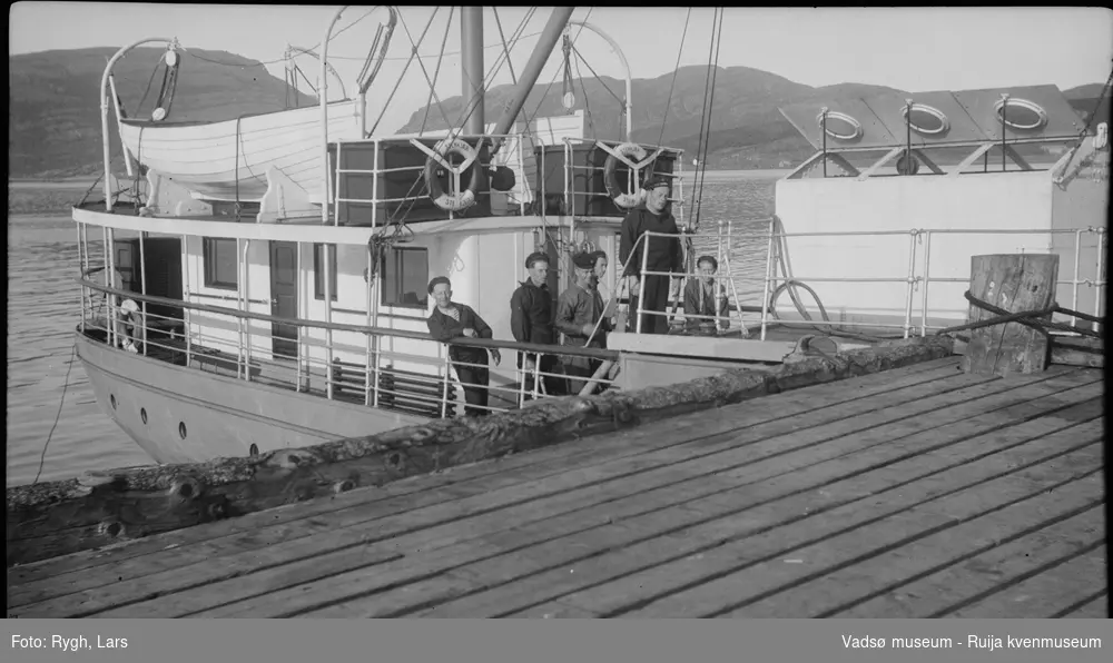 En dampbåt som ligger ved en trekai. På dekket står det seks sjømenn. I bakgrunnen er det fjell, og noen bygninger kan skimtes. Bildet har tilhørt Lars Rygh fra Hønefoss, som var med på minerydding i Varangerfjorden i tidsrommet 1917 - 1918.