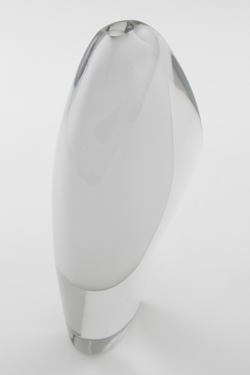Skiveformet vase med en abstrakt, skulpturell utforming i underfangsglass. Det innerste sjiktet består av opakt melkehvitt glass som danner et tilnærmet elipseformet hulerom, mens det ytre sjiktet er i klart glass.