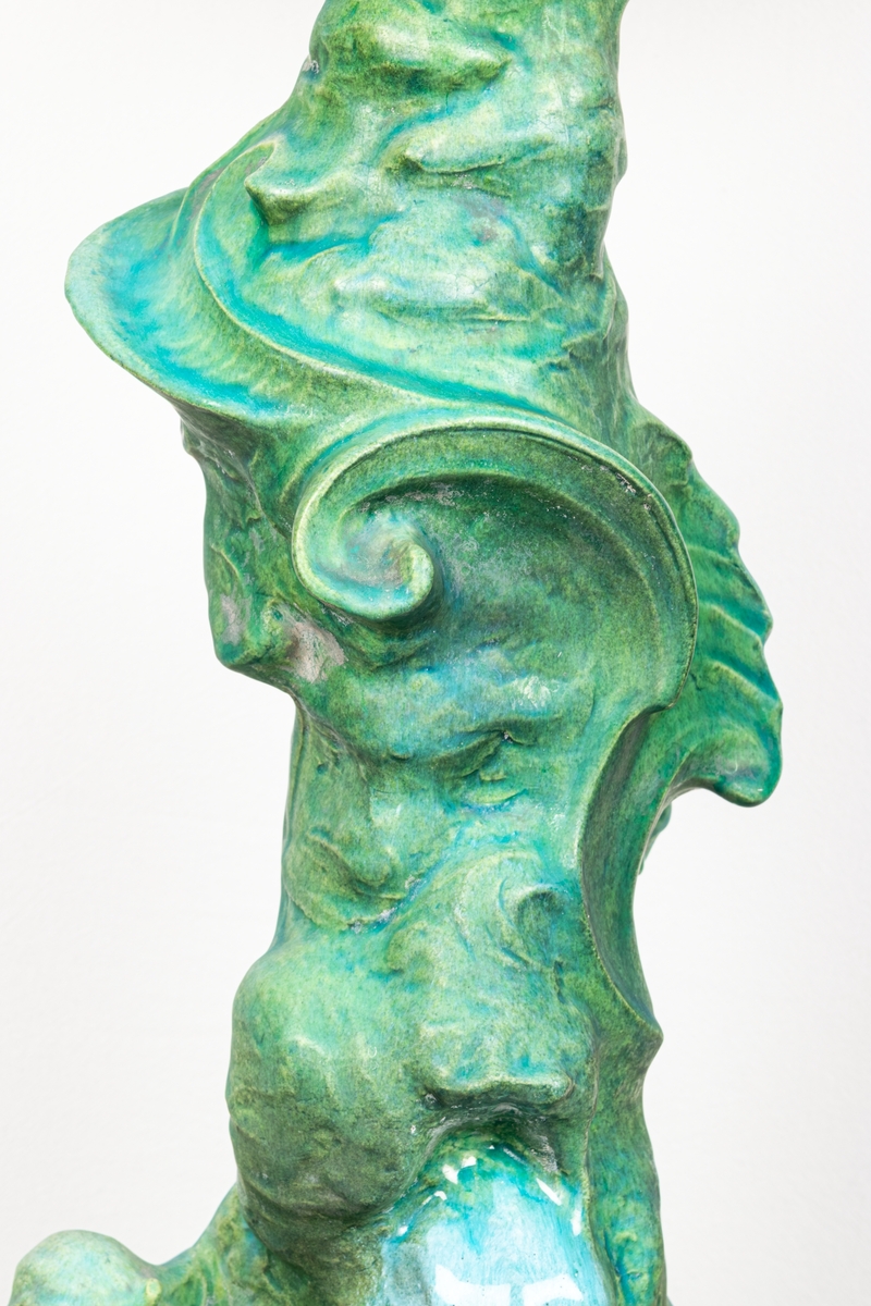Bordsuppsats i keramik med grön glasyr, formgiven här i lera av den franske keramikern Edmond Lachenal efter en originalskiss i gips av Ida Matton, se XLM.42118. Tillverkad i Paris. Föreställer näcken spelande på harpa vid ett vattenfall.