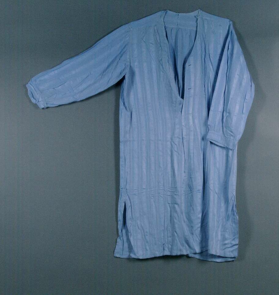 Nattskjorta av ljusblått blandat konstfibermaterial. Vävt med randig struktur med smala vita ränder. V-ringad utan krage. Knäppning framtill med två stora vita knappar.
Söndrig vid halslinningen. Nött.