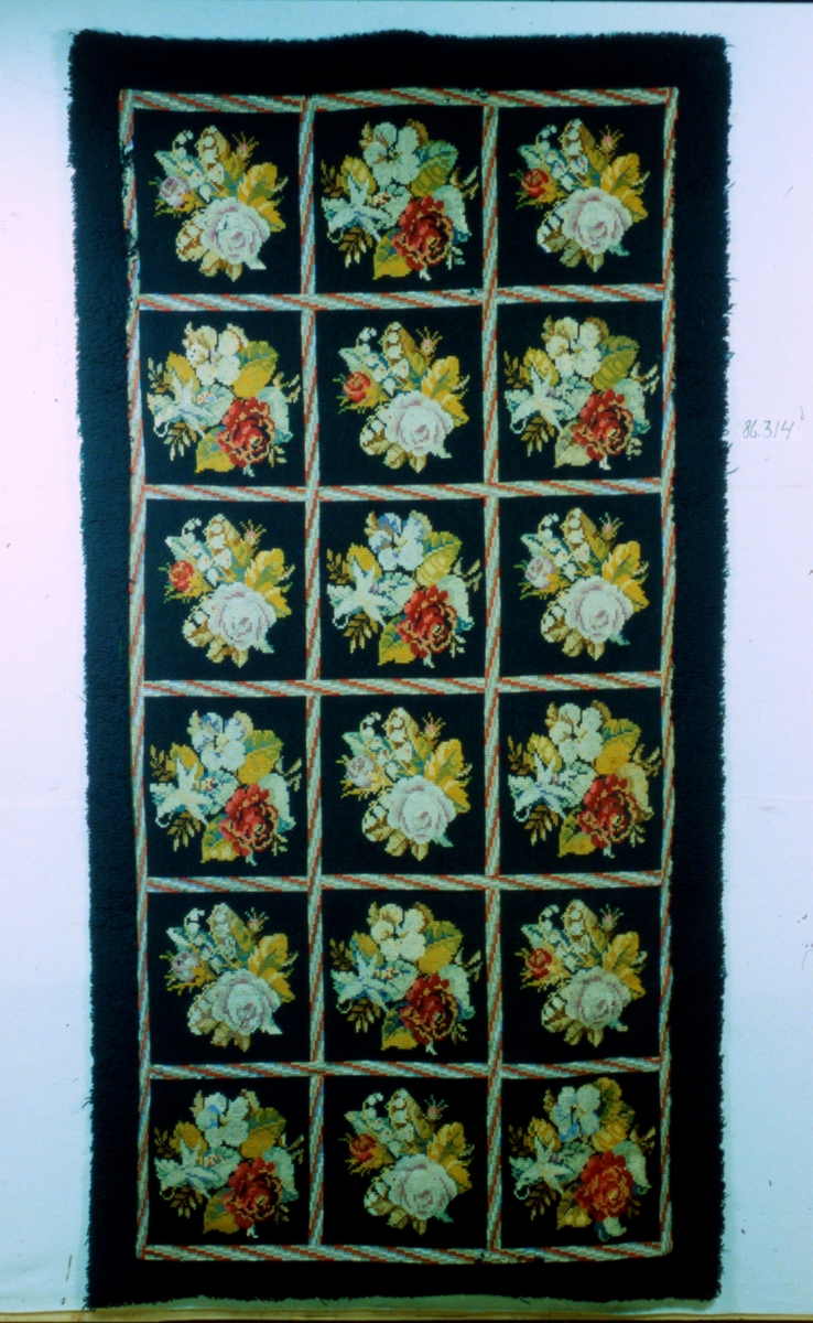 Matta av ylle med korsstygnsbroderi, blommotiv i rutor i många färger på svart botten. ryakant i svart runtom. Svart bomullsfoder. Syddes till fru von Halls bröllop omkring år 1880.