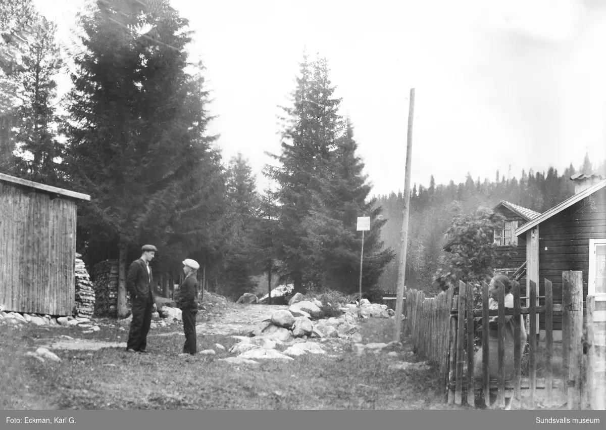 Två pojkar står och samtalar på ett gårdstun, kvinnan bakom staketet invid stugan till höger är Märta Camel. På en fyrkantig skylt i mitten av bilden står det: Motortrafik förbjuden. Ur Karl Eckmans samling.