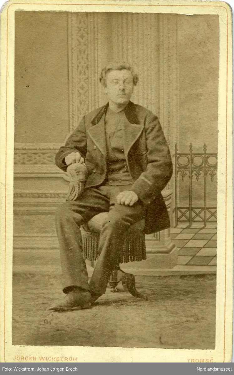 Portrettbilde av en mann sittende på en krakk i klunkerstil, foran malt lerret. Fotoatelier. Peder Olsen, Skånland.