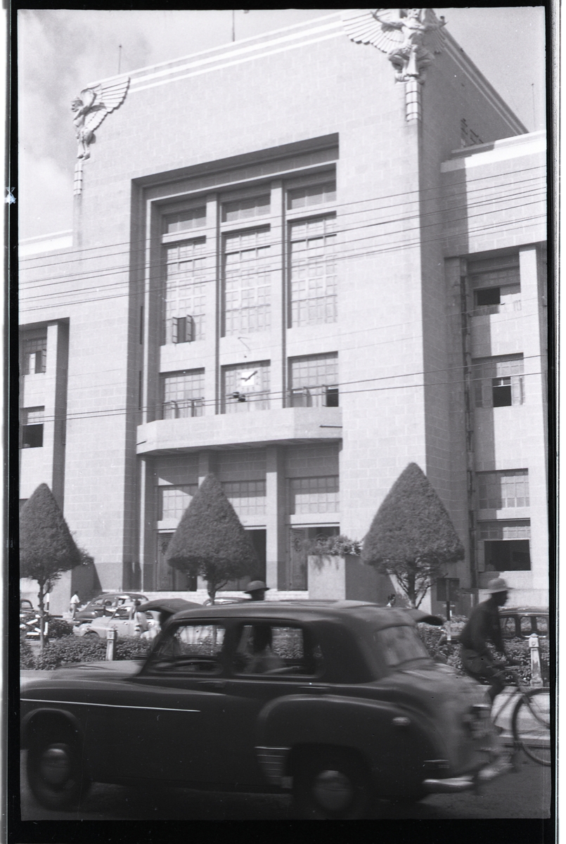 Gatebilde med bil av modell Isuzu Hillman Minx. Bygning utsmykket med griffer (?).
