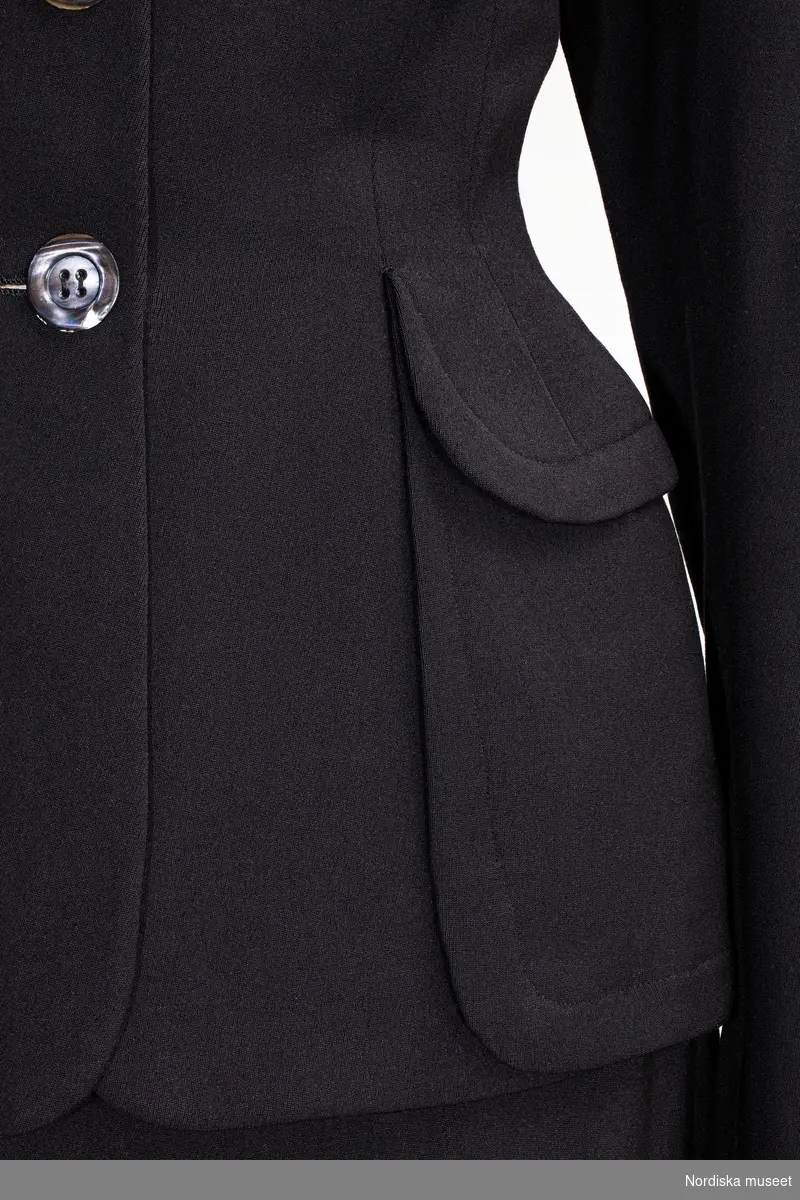 Ylledräkt i figurnära snitt, beställd med två kjolar för variationsmöjligheternas skull och för att kjolen nöttes snabbare. Beställd från NK:s Franska damskrädderi av Margit Graffman (1900-1991) chef för NK:s konfektionsavdelning 1931 till 1953. Levererad juni 1948.
