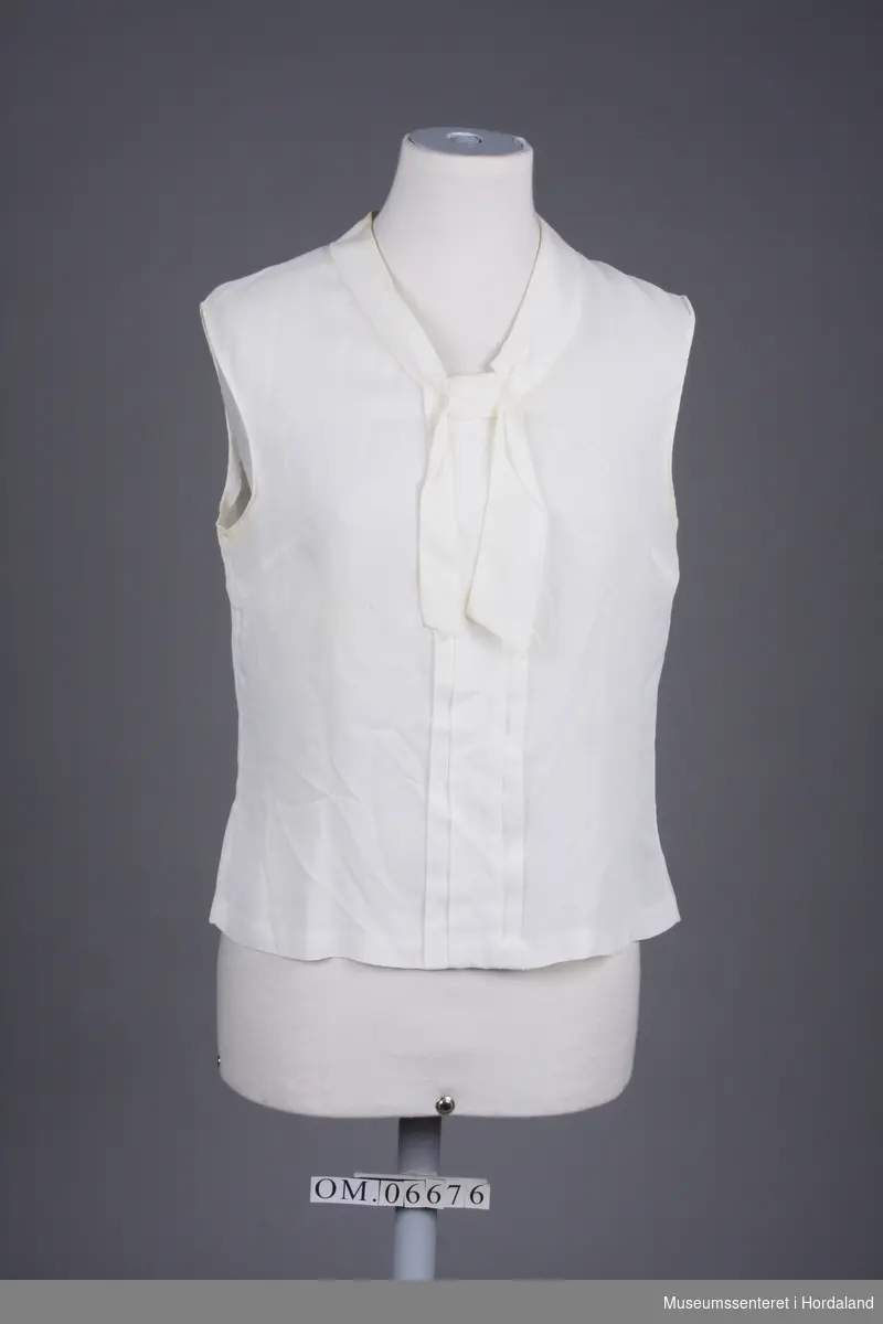 Bluse uten ermer, knappa bak (7 knappar) sløyfe framme i halsen og lagde folder midt framme.
