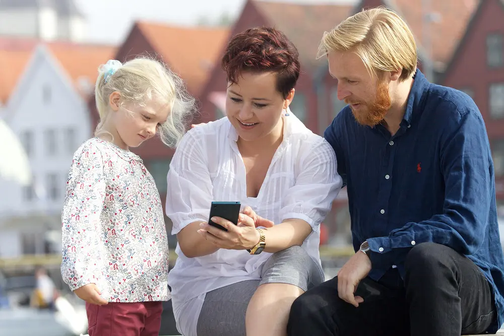 En familie på 3 som ser på mobilen sin og bruker maritim vandringsapp med Bergen Brygge i bakgrunnen.