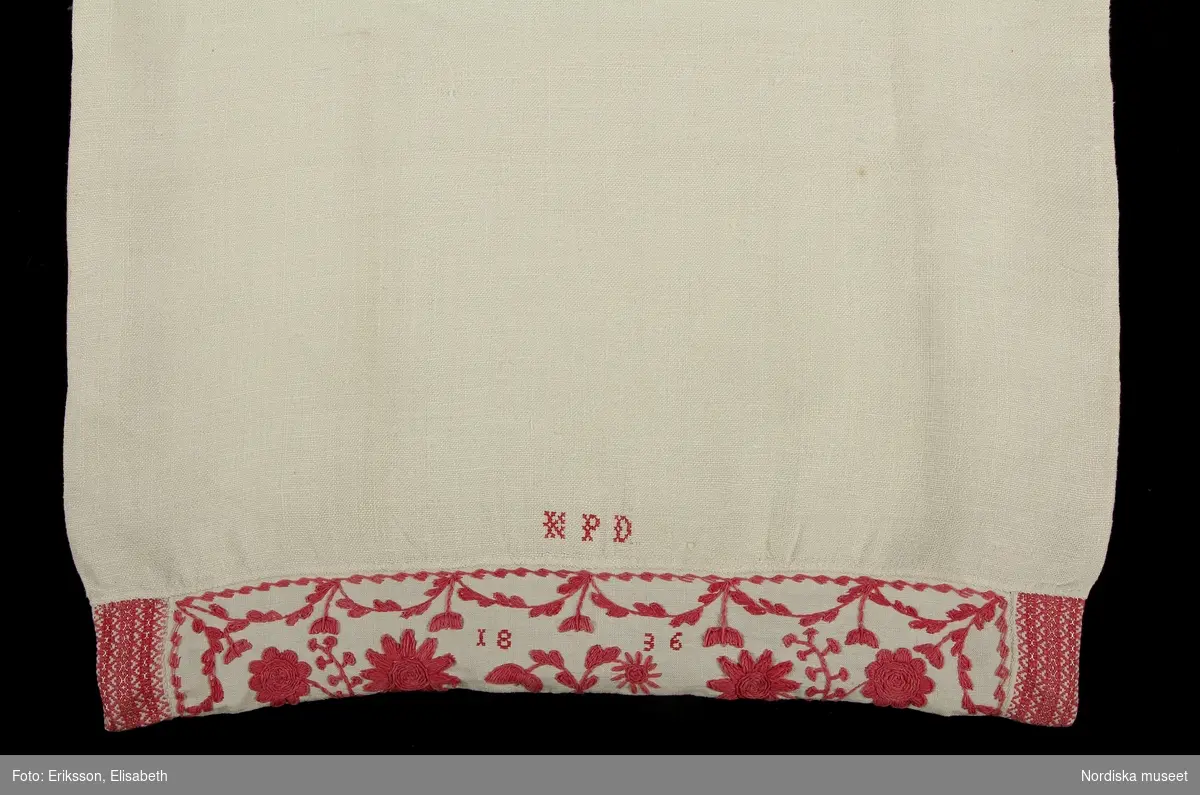 Långörngott av vit linnelärft med fastsydd huva av finare linne med blombroderi med rosarött bomullsgarn, s.k. "turskiskt garn" i plattsöm med konturer i stjälksöm. Ytterst i huvans ändar iskarvade bitar av rosengångsväv i rött och vitt bomullsgarn med stråk av gult silke. Märkt ovanför huvan K P D och inne i broderiet 1836 i korssöm.

Berit Eldvik april 2005