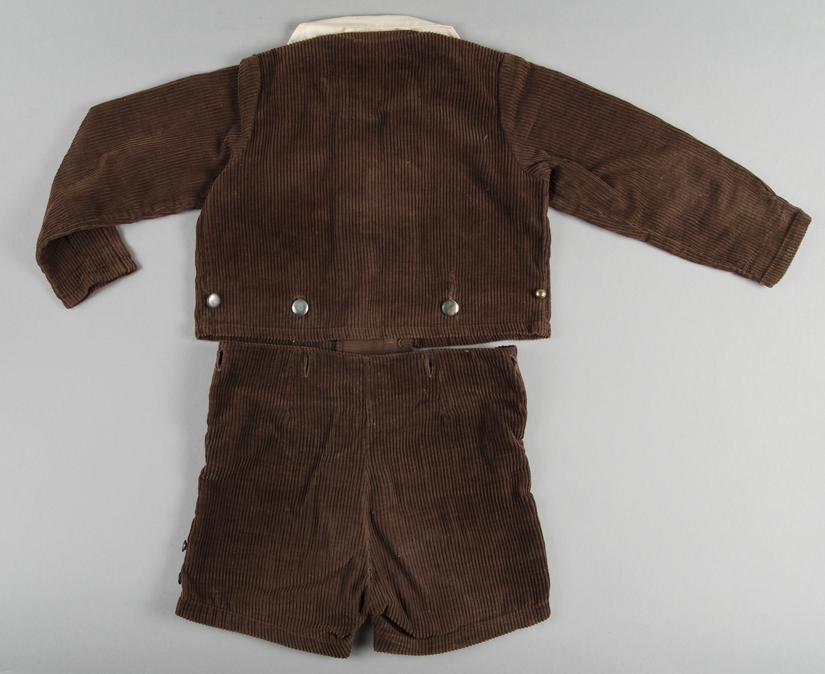 Dräkt för pojke ca 3-5 år. 1920-30-tal. Jacka och kortbyxor i brun manschester. Jackan har lång ärm, knäppes fram med sex blanka metallknappar. I nederkanten av jackan finns ytterligare knappar att fästa byxorna vid. Dräktjackan har löstagbara manchetter, krage och näsduk av gulnat bomulltyg. Bröstficka för näsduk på vänster sida. 
Kortbyxorna knäppes i sidan med runda metallknappar. Knapphål i byxlinningen för för att fästa jackan.

