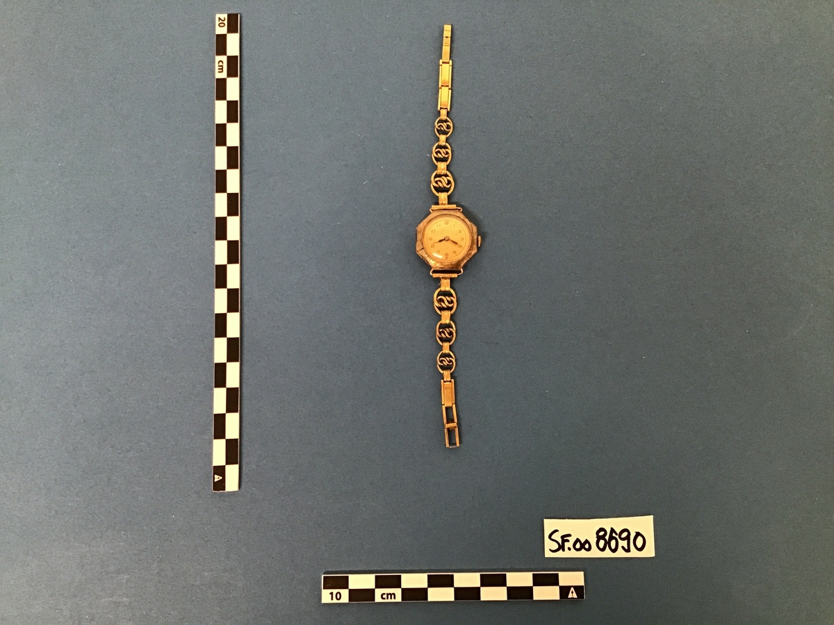 Armbandsur, dameur, i metall, urskiva er kvit med arabiske tegn for markering av 5-minuttsintervall, stigande frå 1 - 12. Metallreim. Sjølve urkassa er åttekanta - oktogon.
