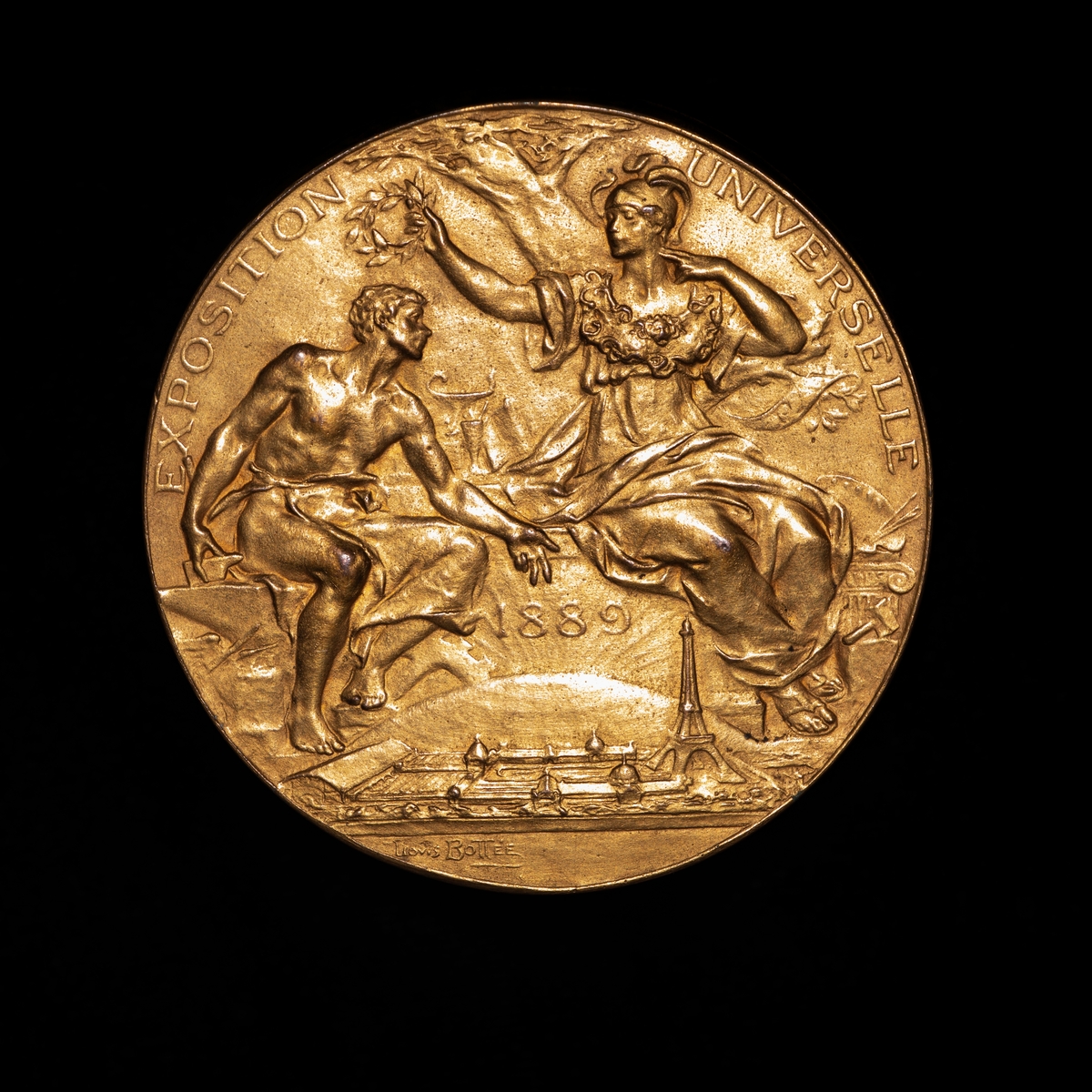 Gullmedalje fra utstilling i Paris 1889. På adversen kvinnelig guddom overrekker laurbærkrans til mann. På revers sittende engel spiller basun.