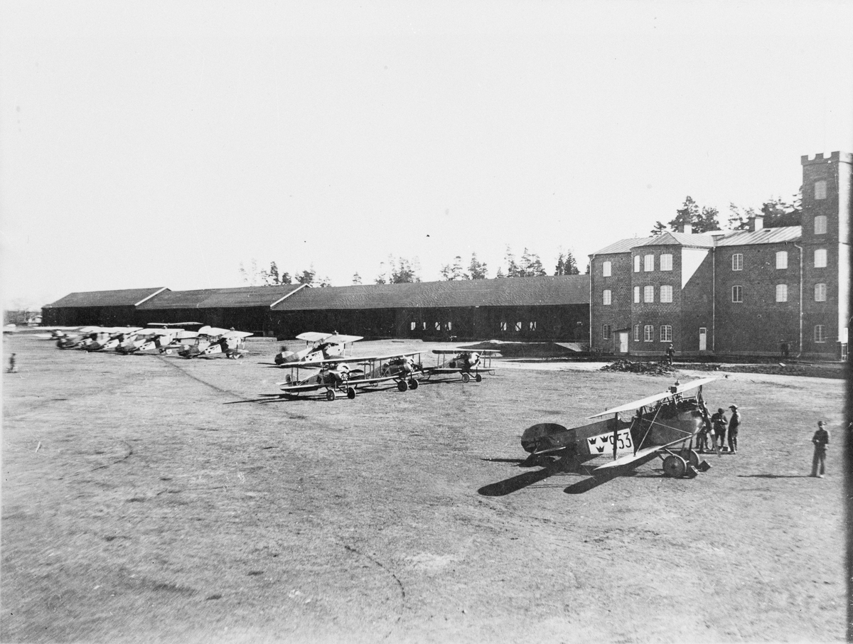 Flygplan framför Flygkompaniet och Centrala flygverkstäderna Malmen. Till höger kanslihuset. Flygplan Phönix D.III nummer 953 i förgrunden, flygplan Tummeliten i mitten och flygplan Phönixjagaren D.II i bakgrunden.