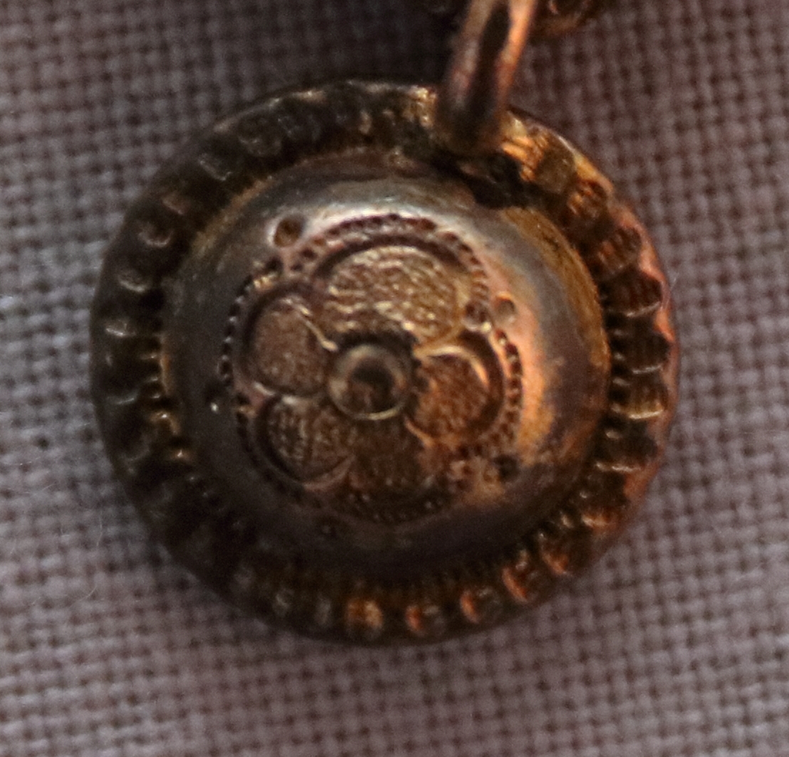 Hängsmycke i förgyllt silver bestående av en slät bas där ovanpåliggande slingrande 'band' av silver är fastsatta. På en rund upphöjning med inristad och pressad dekor (upptill på framsidan av smycket) är en pärla infattad. Nedtill på smycket finns (fastsatt i en hängande ring) en kula med inristad och pressad dekor i form av en blomma.