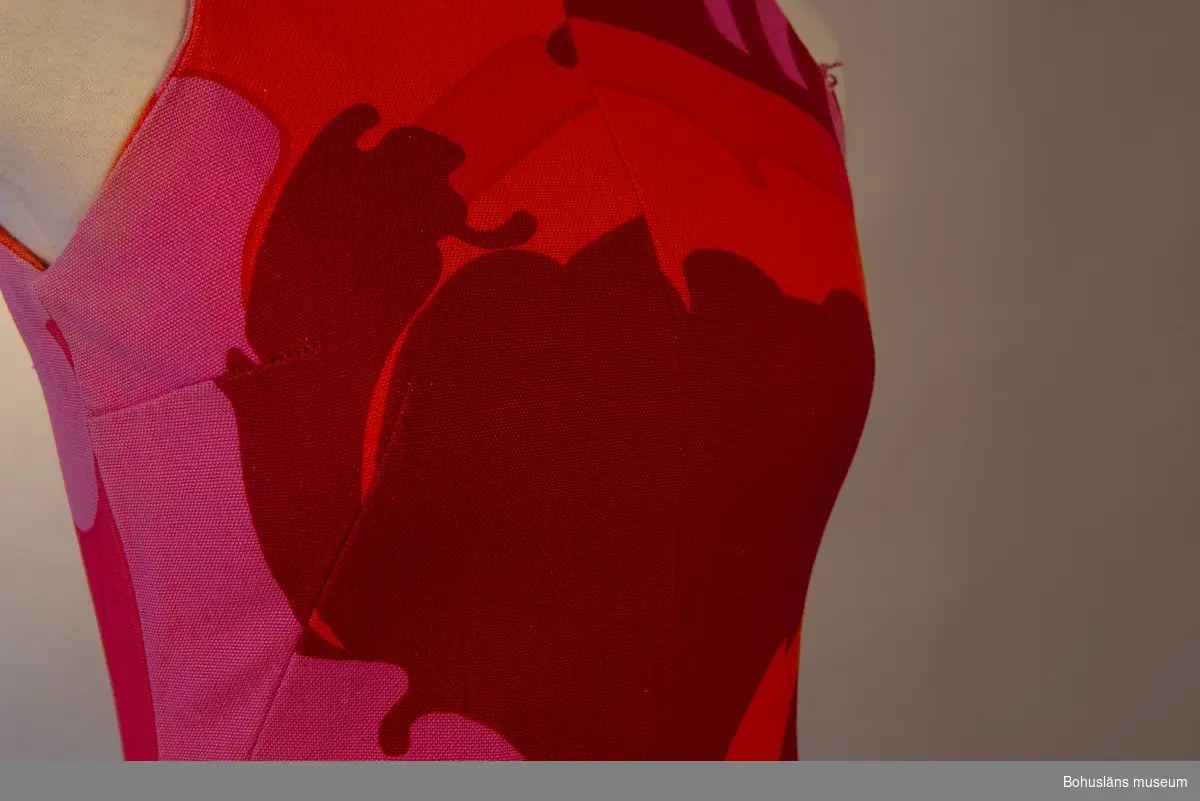 Stormönstrad långklänning i styvt bomullstyg med röd botten. Rak passorm med något utsvängande kjolparti. Sydd av fyra avlånga våder.