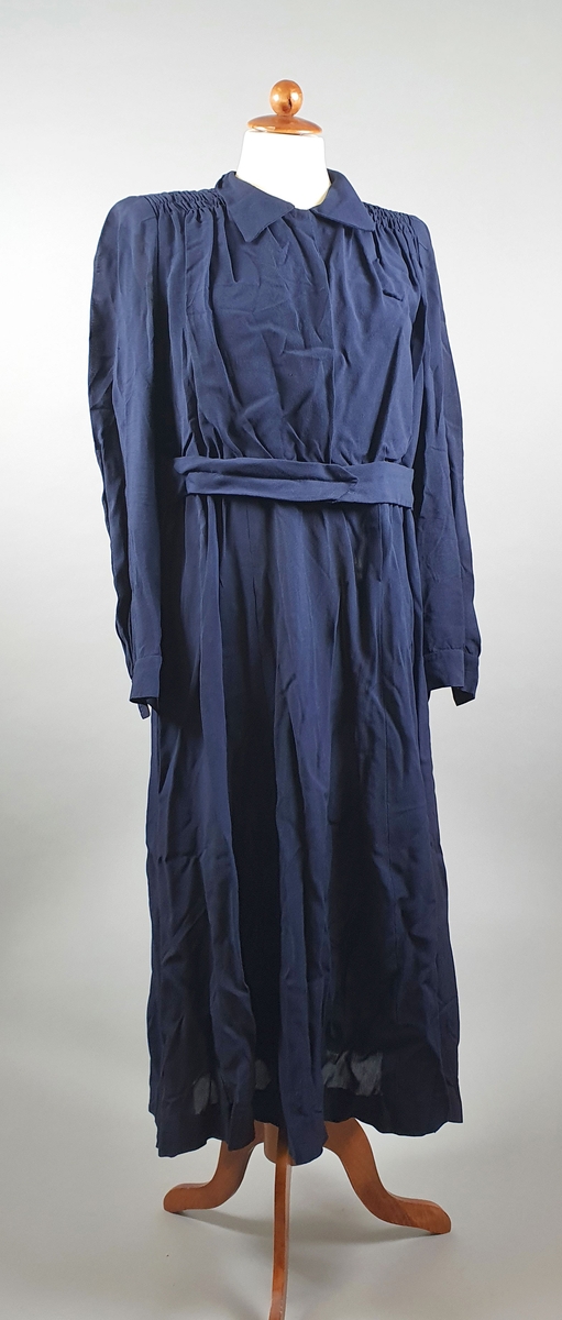 Mørkeblå kjole med lange ermer, skuldreputer, skjortekrage og belte i livet. Kjolen lukkes foran med trykknapper. På hver skulder er stoffet rynket sammen.
