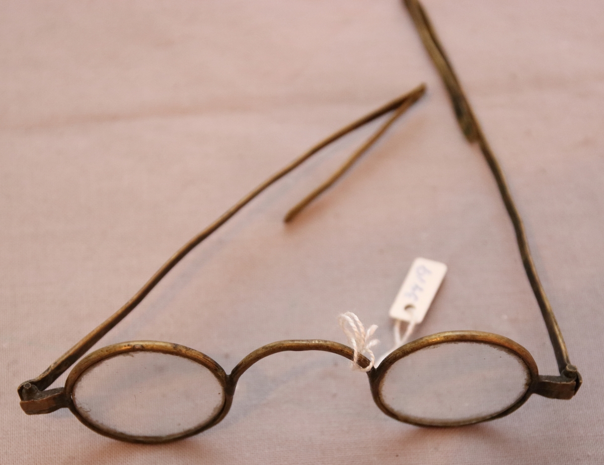 Glasögon i fodral. Fodralet består av två halvor i solida träbitar som sedan är urgröpta så att glasögonen får plats och har gångjärn i metall. Glasögonen har bågar av mässing och runda glas med utfällbara skalmar.