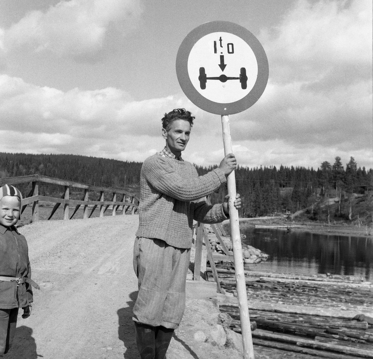 Mann og gutt ved ei veibro, trolig i sørenden av Osensøen i Trysil, Hedmark. Mannen viser et trafikkskilt, høyest tillatt akseltrykk ved passering av brua var ett tonn.