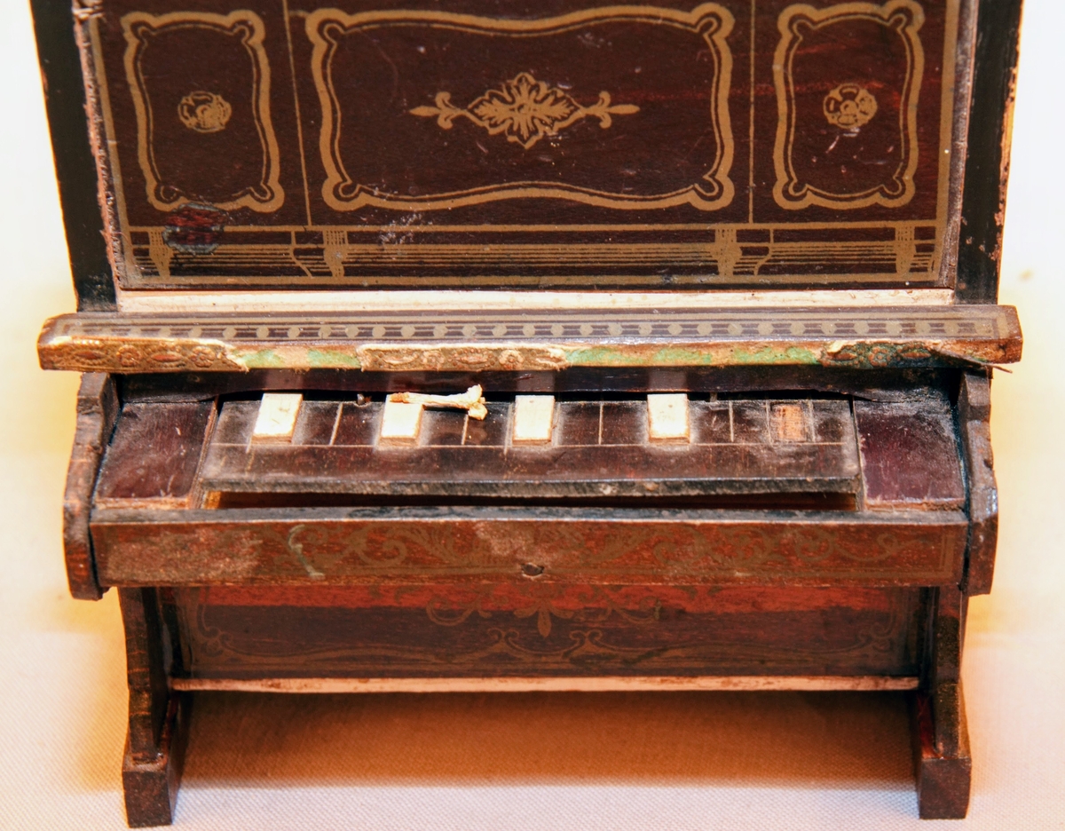 Piano i miniatyr till dockskåp från mitten av 1800-talet. I mörkbrunt målat trä med tryckt dekor i guldig färg på ovansidan och framsidan av pianots ovanstycke och främre nedanstycke. Dekoren föreställer bland annat rocailler, paneler och bårdar i sirlig och asymeterisk stil. Det finns en guldig bård med blomformade maskaroner framtill ovanför tangenterna. Vita fält längst upp och längst ned på främre ovanstycket och nedanstycket (ovanför den tryckta dekoren). Med 15 tangenter (en saknas).