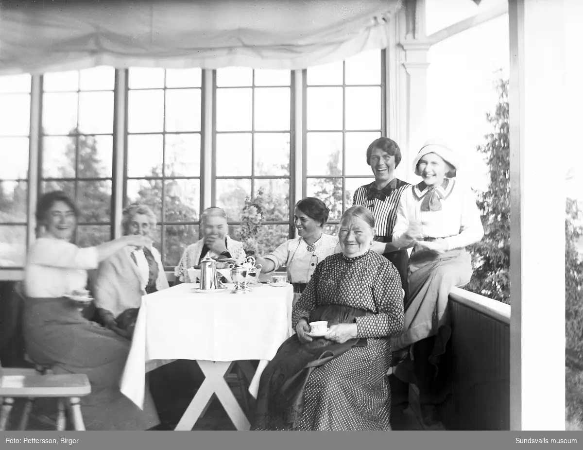 Sju glada kvinnor vid kaffebordet på en glasveranda.