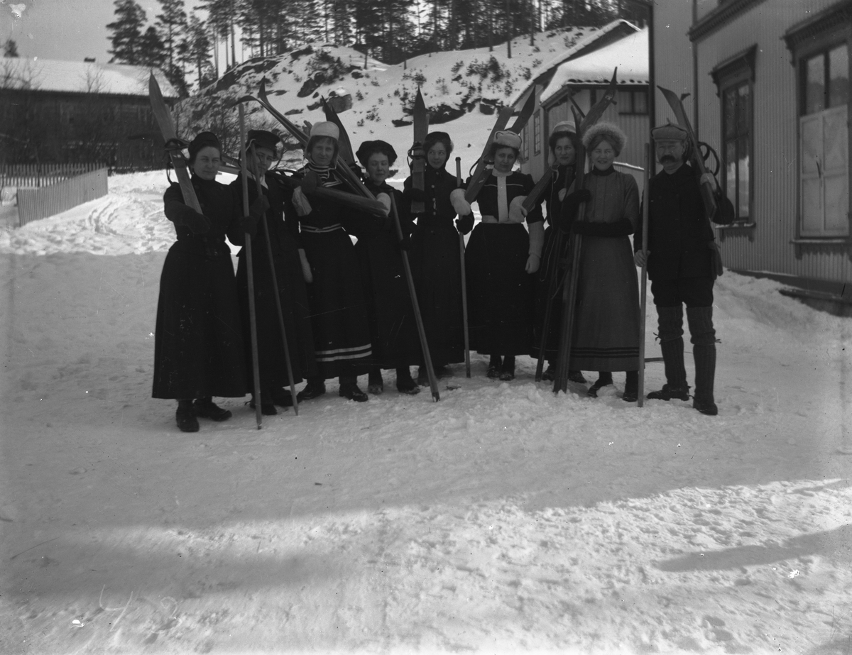Åtte kvinner og en mann med skiutstyr, hus i bakgr.
