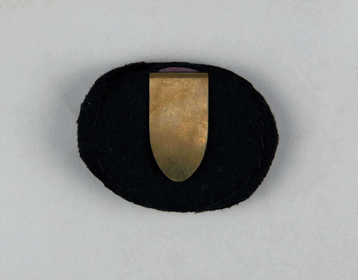 Ovalt mössmärke av metall, klädd med svart tyg med broderat vinghjul av metalltråd och nationalkokard av tyg. Metallspänne på baksidan för fastsättning.
