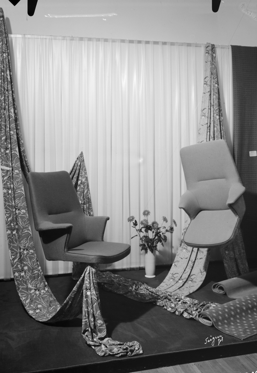 Utstillingen "Norsk tekstil 1952" med stoler trukket med vevde møbelstoffer, trykte og gjennomskinnelige gardiner og teppeprøver. I midten er dekorativ blomsterbukett.