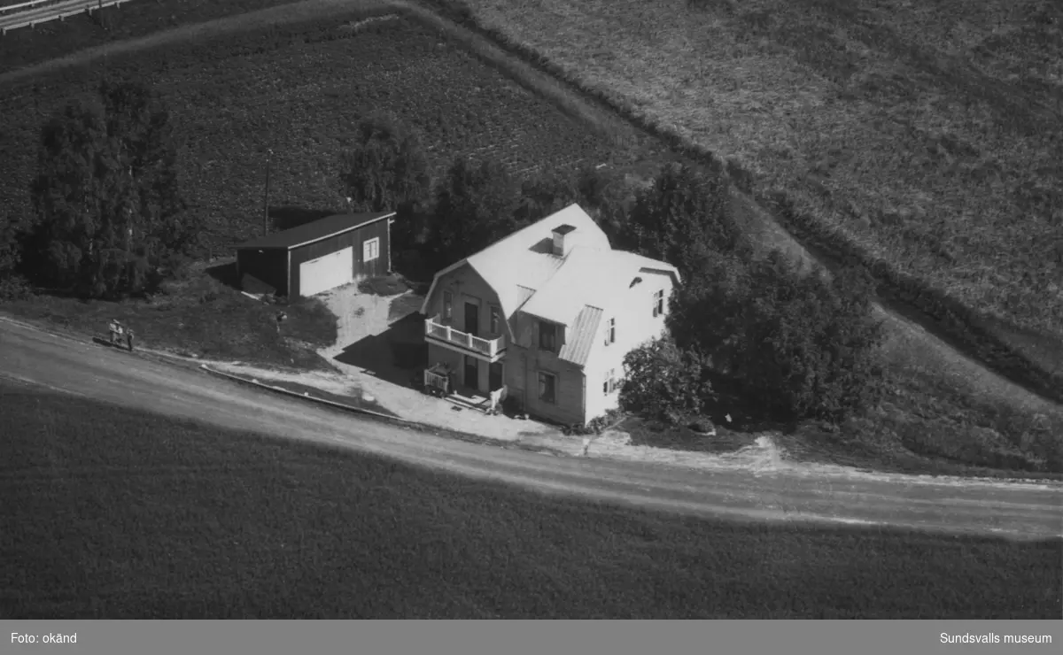 Flygfoto över fastigheten Strömås 1:10. Vid vägkanten syns en kvinna och två barn. Ur en samling som tillhört Lotta Nordlund, Strömås.