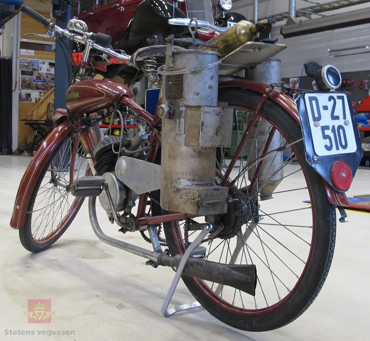 Rød (vinrød), motorsykkel med pedaler, forbrenningsmotor og vedgassgenerator. 
Motorsykkelen er en BLIXT lettvekts motorsykkel fra 1936 med en 98 cc, en-sylindret totaktsmotor av fabrikat Fichtel & Sachs som yter 2,5 hk og går på bensin.
Generatoren er av fabrikat Åsbrink, type Sperco lI, og laget primært for bruk med trekull. Virkningsgraden er ca 0,4 med "godt køl", det vil si en 2,5 hk motor kan under gunstige omstendigheter teoretisk utvikle ca 1 hk. Kullet skal knuses til biter av størrelse 5-15 millimeter, og oppfyringen er svært omstendelig. I teorien skal man kunne kjøre ca 10 km på "en fyring".
Motorsykkelen har 2 gir og veier 73 kilo.
Den er 72 cm bred, 210 cm lang, og ...høy...
På hele sykkelen er det 1 sitteplass.
Det er montert pumpe til dekkene på ramma og et brannslukkingsapparat bak på sykkelen.