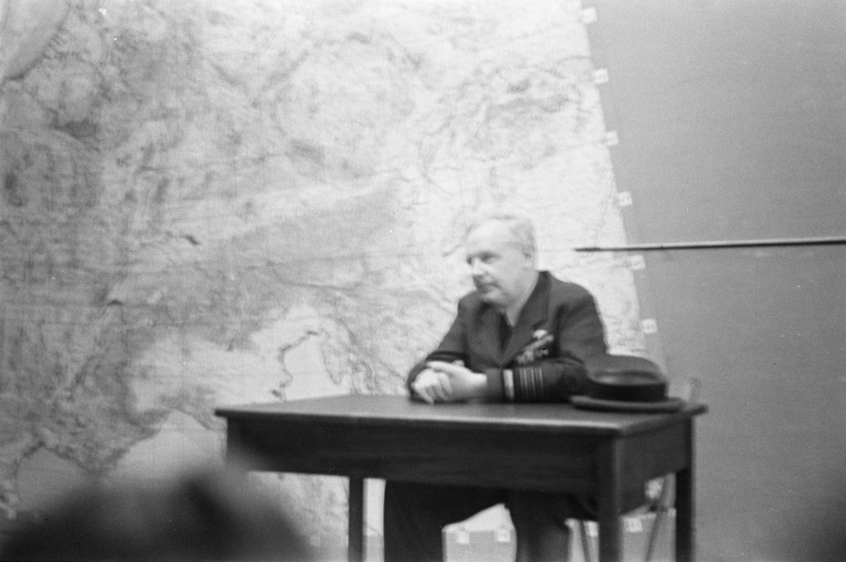 Person i uniform sitter ved bord foran stort kart uskarpt bilde