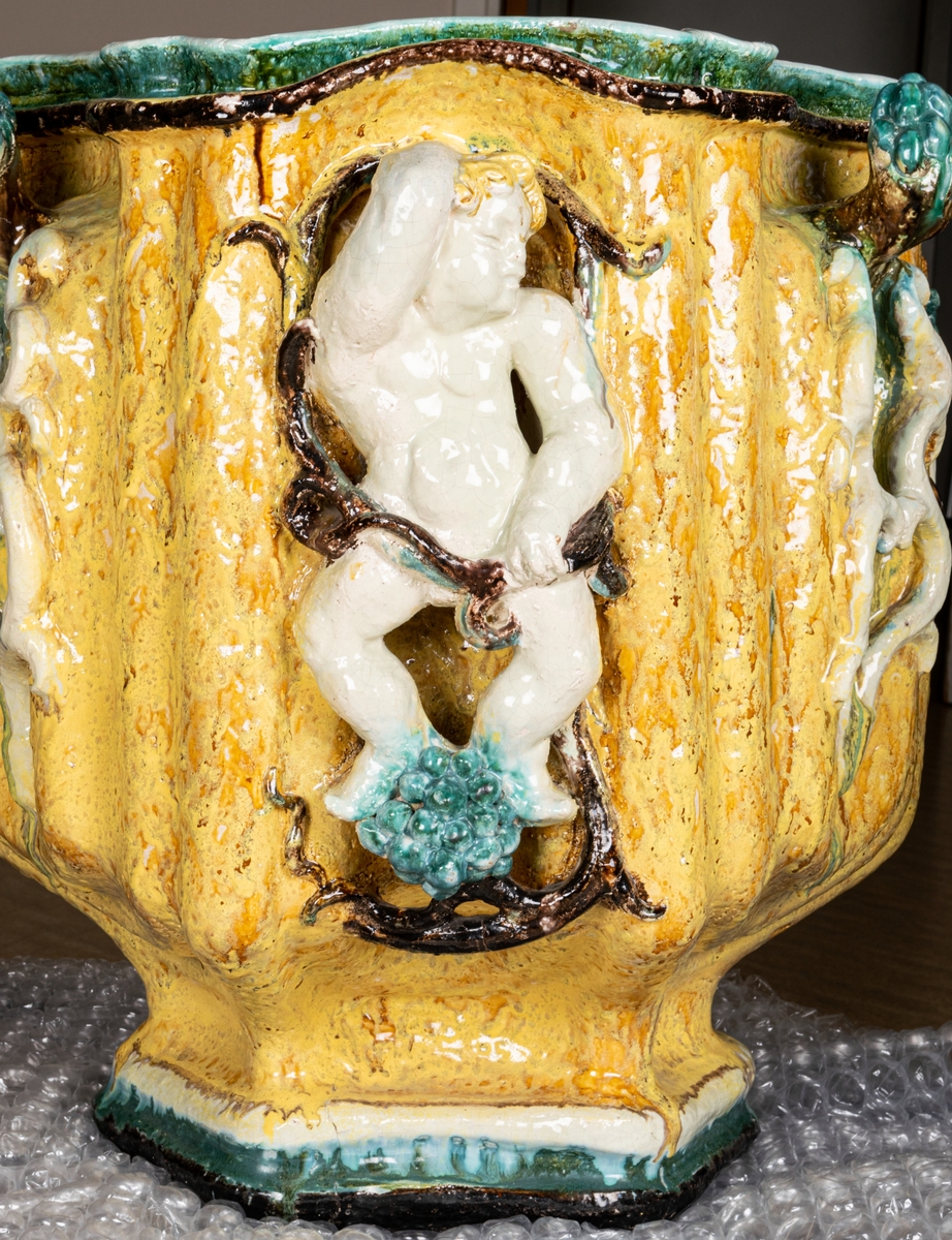 Terassurna, Bo Fajans, design Allan Ebeling. Dekor av skulpterade amoriner med frukter i vitt, grönt, blått och brunt mot gul antimonglasyr. Vågig överkant, 6-sidig fot.