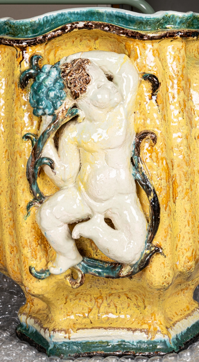 Terassurna, Bo Fajans, design Allan Ebeling. Dekor av skulpterade amoriner med frukter i vitt, grönt, blått och brunt mot gul antimonglasyr. Vågig överkant, 6-sidig fot.