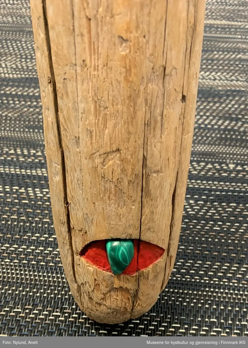 Lang rekved, med delvis rød og mørkgrønn farge. En fjær settet fast. Helt nederst er det laget et øye av rød farge og en grønn malakitt.